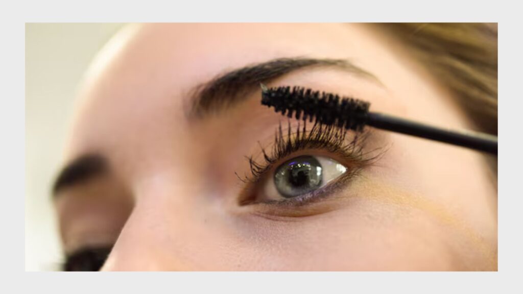 A close-up of a woman applying mascara or eyelash serum to her lashes with a brush