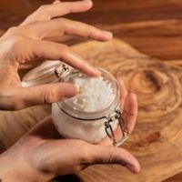 A person holding a small glass jar of salt or scrub against a wooden surface