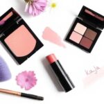 Kaja Dewy Blush and Lip Balm Duo on a white background, with soft lighting, makeup swatches, and natural props like flowers and brushes – beauty flat lay